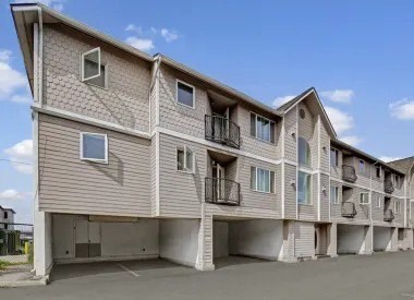 Three-story apartment building here at La Maisonnette with beige siding and white trim. The building features private balconies, covered parking beneath the structure, and multiple exterior windows in a residential setting.