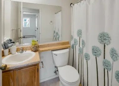 A bathroom here at La Maisonnette with a bathtub-shower combo and a patterned shower curtain. The space features a white toilet, a wooden vanity with a beige countertop, a single sink, and a wall mirror, offering a clean and functional layout.