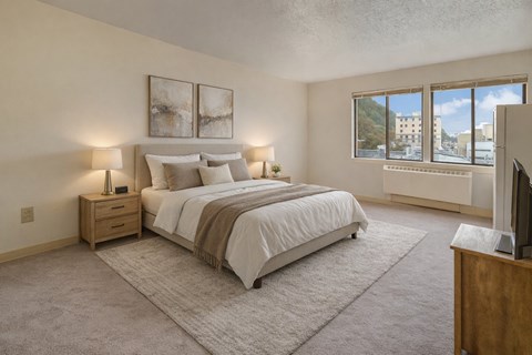 A bright, cozy bedroom here at Mendenhall-Tower with soft neutral carpet, a comfortable bed dressed in white linens, and warm bedside lighting. A large window brings in natural light and scenic city views, creating an airy and relaxing atmosphere.