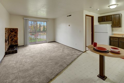 A spacious living room with a dining table and chairs, a carpeted floor, and a view of a parking lot through a sliding glass door.