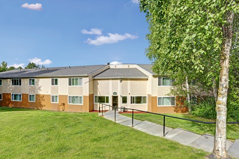 A building with a brown and beige exterior is surrounded by a green lawn and trees.