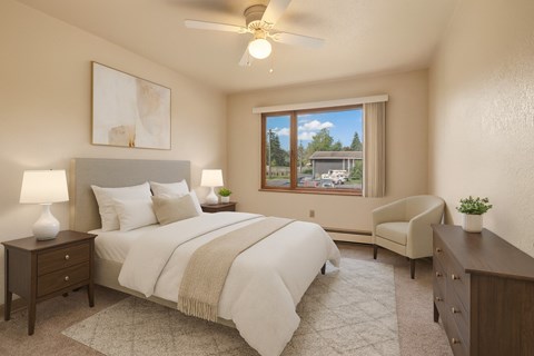 A bright bedroom at The Alaskan with soft carpeting and warm neutral walls. The space features a neatly made bed with matching bedding, two bedside tables with lamps, and a dresser. A large window brings in natural light and offers views of the surrounding neighborhood.