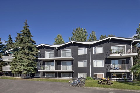 A three-story exterior view of The Alaskan with dark siding, white trim, and private balconies on each level. Mature evergreens and open lawn space create a natural setting, with a picnic table and bicycles adding to the community-friendly atmosphere under a bright blue sky.