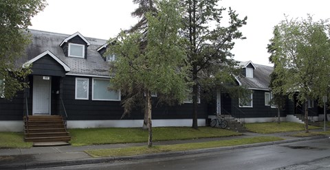 A long single-story building at The Alaskan with dark siding, white trim, and multiple front entrances accessed by small stairways. Mature trees line the sidewalk and grassy lawn, creating a residential neighborhood feel.