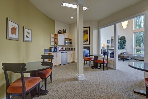 A cozy resident café area at The Alpine featuring small round tables with cushioned seating, a beverage station with a microwave, coffee maker, and mini fridge, and open shelving for supplies. Warm earth tones, framed artwork, and carpeted floors create a relaxed space next to the building’s lobby.