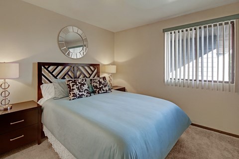 A cozy bedroom at The Alpine featuring a neatly made bed with blue bedding and patterned accent pillows. Matching bedside tables with lamps sit on each side of the headboard, and a window with vertical blinds brings in natural light. A round wall mirror adds a stylish touch above the bed.