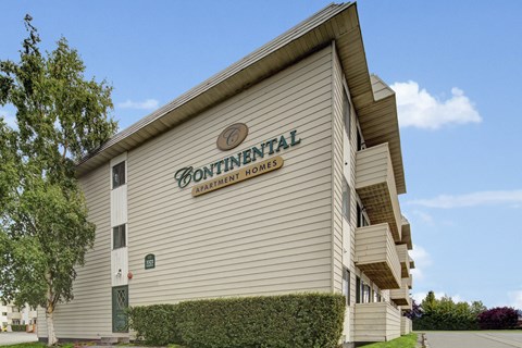 A bright exterior view here at The Continental featuring beige apartment buildings with multiple balconies, clear Continental Apartment Homes signage on the facade, manicured hedges, mature trees, and a paved driveway under a blue sky.