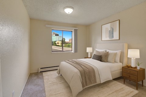 A serene bedroom here at The Continental featuring a queen-sized bed with white bedding and a soft neutral throw, matching bedside tables with lamps, neutral carpet flooring, abstract wall art, and a large window that brings in natural light and neighborhood views.