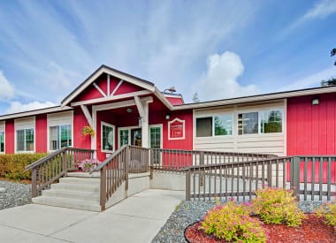 A bright community building exterior here at The Glen featuring a welcoming red-and-neutral facade with white trim, a central gabled entrance, and a fully accessible ramp and stairs leading to the front doors, surrounded by neatly landscaped beds, decorative gravel, and colorful shrubs under a partly cloudy blue sky.