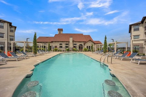 A resort-style outdoor swimming pool here at Palisades at Pleasant Crossing featuring a long rectangular pool with clear blue water, built-in water features, surrounding lounge chairs with shaded pergolas, wide concrete deck space, and a stylish clubhouse backdrop under a bright blue sky.