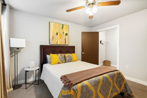 A cozy, well-lit bedroom here at Palisades at Pleasant Crossing featuring a neatly made bed with neutral bedding and warm yellow accent pillows, a padded headboard, modern abstract wall art, soft carpet flooring, a ceiling fan with light, and an open closet that adds functional storage to the inviting space.