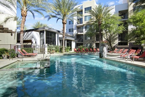 A resort-style swimming pool here at Acclaim featuring clear blue water, surrounding palm trees, red lounge chairs, shaded seating areas, and modern apartment buildings that create a relaxing and inviting outdoor community setting.