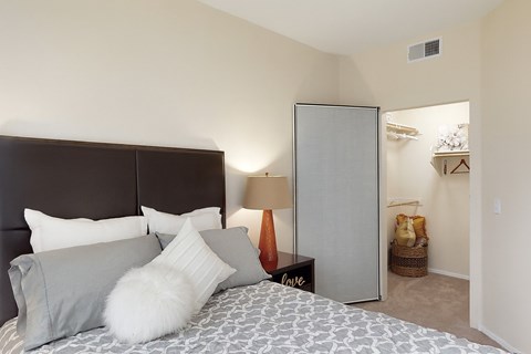 A cozy bedroom here at Acclaim featuring a plush bed with neutral bedding and layered accent pillows, a padded headboard, a bedside table with warm lamp lighting, soft beige carpeting, and an open walk-in closet with shelving and hanging space that adds everyday functionality.