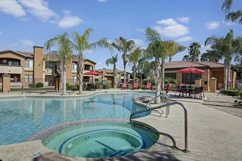 A resort-style outdoor pool here at Arezzo featuring a curved swimming pool with an attached spa, palm trees, red lounge chairs, shaded seating areas, and surrounding apartment buildings that create a relaxing, sunny setting ideal for cooling off, socializing, or enjoying the landscaped community courtyard.