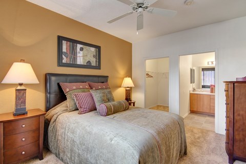 A cozy primary bedroom here at Arezzo featuring soft carpeted flooring, a comfortable bed with layered bedding and accent pillows, warm-toned accent walls, matching nightstands with lamps, a ceiling fan for added comfort, and direct access to a spacious walk-in closet and an adjoining bathroom with a wide vanity.