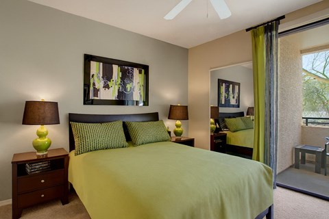 A stylish bedroom here at Aspire Pinnacle Park featuring a plush bed dressed in vibrant green bedding with coordinating patterned pillows and rich wood nightstands on either side. Matching table lamps add warm lighting, while abstract wall art and a mirrored closet reflect natural light from the sliding glass door leading to a private patio.