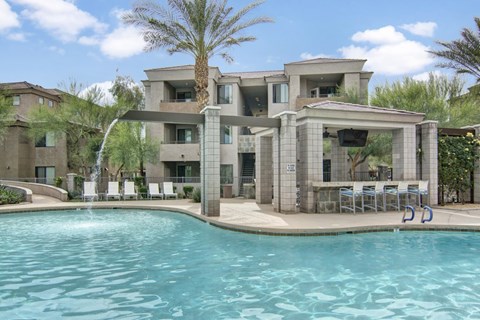 A resort-style swimming pool here at Aspire Pinnacle Park featuring sparkling turquoise water, a graceful fountain feature, and a shaded cabana with bar seating and mounted television. Lounge chairs line the expansive sundeck beneath tall palm trees, while surrounding apartment buildings create a private, desert-inspired retreat perfect for relaxing afternoons or social gatherings.