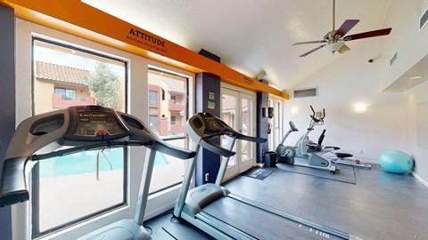 A bright fitness center here at Aspire Pinnacle Park featuring treadmills, elliptical machines, and cardio equipment positioned near large windows overlooking the pool area. Vaulted ceilings with a ceiling fan, motivational wall accents, and durable flooring create an energizing space designed for convenient, comfortable workouts close to home.