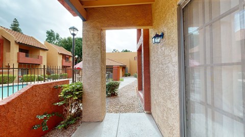 A charming covered patio here at Aspire Pinnacle Park featuring textured stucco walls, a private entryway, and a sliding glass door that brings in natural light. The walkway overlooks landscaped grounds and a sparkling pool area, offering a cozy outdoor space to relax while enjoying the community’s inviting courtyard setting.