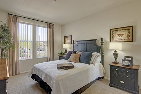 A bright and inviting bedroom here at Bela Rosa featuring a dark wood bed frame with layered neutral bedding and accent pillows. Large windows with vertical blinds and drapery panels fill the room with natural light, while matching nightstands with decorative lamps and framed artwork create a warm, cohesive retreat with plush carpeting underfoot.