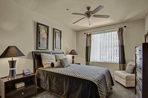 A serene and inviting bedroom here at Bolero featuring a spacious layout with plush carpeting, a sleek dark wood bed frame, and coordinating nightstands with modern table lamps. Large windows with vertical blinds and accent curtains fill the space with natural light, while a ceiling fan enhances comfort and the cozy seating nook adds a relaxing retreat feel.