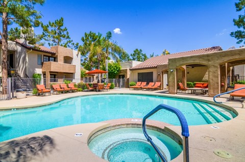A resort-style pool area here at Desert Horizon featuring a sparkling swimming pool with a separate spa and built-in steps. Lounge chairs with bright cushions line the deck, while shaded seating areas and surrounding trees create a relaxing atmosphere. Nearby apartment buildings and landscaped grounds complete the inviting community setting.