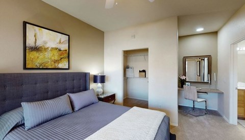 A comfortable bedroom here at Dobson Towne Center featuring plush carpet, neutral walls, and a ceiling fan with light. A spacious bed with an upholstered headboard anchors the room, while an open closet and built-in vanity area add functionality. An adjoining bathroom enhances privacy and everyday convenience.
