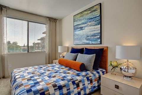A stylish bedroom here at Escape featuring a warm wood headboard, layered blue and gray bedding with an accent bolster pillow, and matching nightstands topped with modern chrome lamps, while a large window with blinds and soft drapery fills the space with natural light and a bold abstract artwork adds a contemporary focal point above the bed.