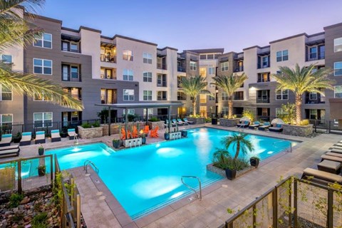 A resort-inspired pool courtyard here at The Flats at San Tan featuring a sparkling turquoise swimming pool with wide entry steps, surrounding lounge chairs with cushioned seating, illuminated palm trees, and a covered cabana with vibrant Adirondack chairs, all framed by contemporary apartment homes and warm evening lighting for a relaxing community retreat.