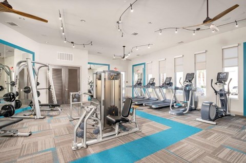 A spacious fitness center here at The Flats at San Tan featuring strength-training machines, free weights, and multiple cardio options including treadmills and elliptical machines, all arranged on patterned flooring with teal accents, large windows for natural light, ceiling fans, and modern lighting for an energizing workout environment.