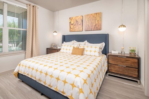 A serene and stylish bedroom here at The Flats at San Tan featuring a plush upholstered bed dressed in white bedding with golden patterned accents, complemented by matching nightstands, warm wood finishes, and modern pendant lighting, while a large window with soft curtains brings in natural light to create a bright and inviting retreat.