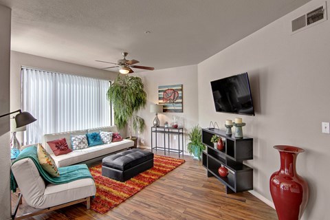 A vibrant living room here at Hawthorne featuring a sleek white sofa with colorful accent pillows, a plush red and orange area rug, and rich wood-style flooring, complemented by a mounted flat-screen television, modern black media console, decorative plants, and a large sliding glass door with vertical blinds that fills the space with natural light.