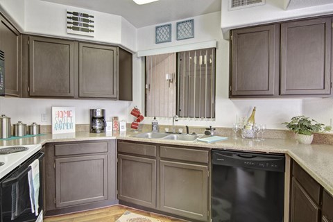 A functional kitchen here at Hawthorne featuring warm brown cabinetry, expansive countertops, and a double-basin sink centered beneath a window with vertical blinds, complemented by a full appliance package and ample prep space, creating an efficient layout that supports everyday cooking, storage, and casual entertaining.
