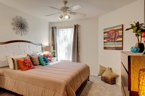 A cozy bedroom here at Hawthorne featuring a plush tufted headboard, soft neutral bedding accented with vibrant decorative pillows, and a ceiling fan with overhead lighting, complemented by a large window with vertical blinds and elegant drapery that invites natural light while maintaining privacy for a relaxing and comfortable retreat.