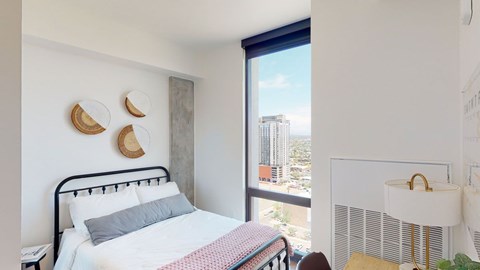 A bright and modern bedroom here at JOY on 4th featuring a sleek black metal bed frame with crisp white bedding and a soft accent throw, complemented by woven wall décor and a concrete accent column. Expansive floor-to-ceiling windows showcase sweeping city views while natural light enhances the clean lines, warm wood flooring, and contemporary finishes.