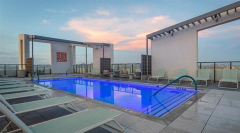 A stunning rooftop pool deck here at JOY on 4th featuring a sleek rectangular swimming pool illuminated with vibrant blue lighting and surrounded by modern lounge chairs. Framed by clean architectural lines and glass railings, this elevated retreat offers sweeping city views, open sky sunsets, and stylish seating areas designed for relaxing, socializing, and soaking in the urban atmosphere.