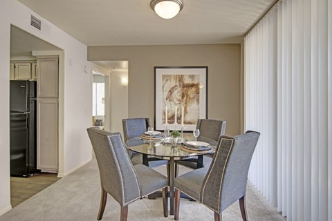 A warm welcoming dining area here at Mirador featuring plush carpet flooring and a round glass-top table surrounded by four upholstered chairs with nailhead trim. A large framed art piece anchors the space, while vertical blinds filter natural light and an open entryway leads toward the kitchen, creating an inviting setting for meals and gatherings.