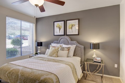 A cozy bedroom here at Mirador featuring a plush upholstered headboard, layered white and gold bedding, and two matching bedside tables with modern lamps. A large window with blinds invites natural light, while soft carpet flooring and neutral walls create a calm, comfortable retreat perfect for rest and relaxation.