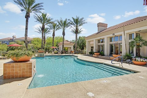 A resort-style swimming pool here at Mirador surrounded by tall palm trees, terracotta tile accents, and spacious sun decks. The sparkling blue water curves alongside wide entry steps and stainless handrails, while vibrant planters, shaded seating areas, and a stucco clubhouse with red tile roofing create a warm, inviting atmosphere perfect for relaxing outdoors.