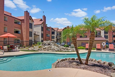 A resort-style pool area here at Motif featuring sparkling blue water, palm trees, and landscaped rock accents. Lounge chairs and shaded patio seating provide space to relax, while the surrounding three-story apartment buildings with balconies create a welcoming community atmosphere under a bright, sunny sky.