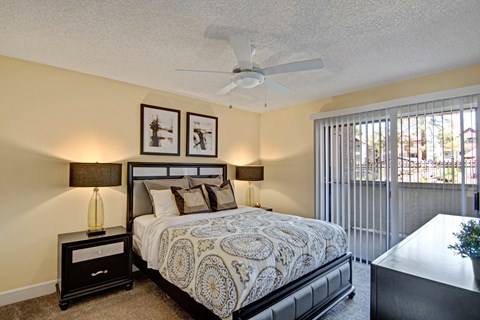 A cozy bedroom here at Motif featuring plush carpet, soft neutral walls, and a ceiling fan with light fixture. A large sliding glass door with vertical blinds opens to a private patio, bringing in natural light, while matching bedside tables and lamps create a balanced, inviting retreat.