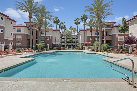 A resort-style swimming pool here at Mozaic at Steele Park surrounded by tall palm trees, comfortable lounge chairs, and shaded patio seating. The sparkling blue water is framed by spacious sundecks and modern apartment buildings, creating a relaxing outdoor retreat perfect for soaking up the Arizona sunshine or enjoying time with neighbors.