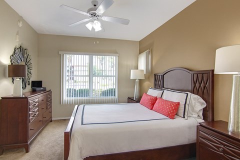 A comfortable bedroom here at Mozaic at Steele Park featuring plush carpet, neutral walls, and a ceiling fan with light. A large window with vertical blinds brings in natural light, while the spacious layout accommodates a full bedroom set including matching nightstands and a dresser for added storage.