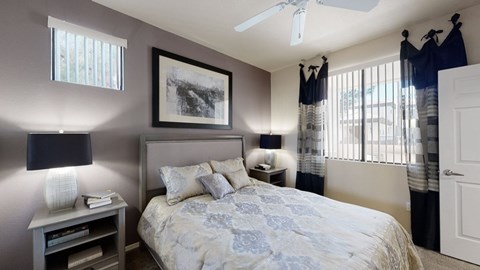 A serene bedroom here at Natura Villas featuring a plush bed with patterned bedding and coordinating accent pillows, framed by soft neutral walls and a contrasting accent wall. Matching bedside tables with modern lamps add balance, while large windows with vertical blinds and layered curtains invite natural light, creating a calm and comfortable retreat.