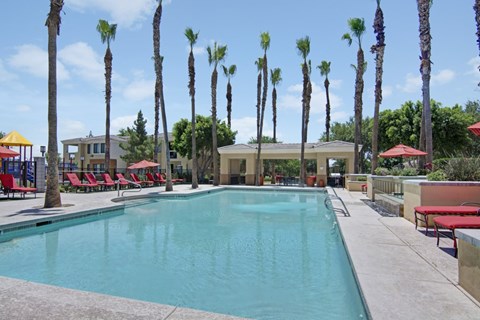 A resort-style swimming pool here at Natura Villas surrounded by tall palm trees and expansive sun decks, featuring crystal-clear water, multiple lounge chairs with vibrant red cushions, and shaded seating areas under umbrellas. A covered pavilion offers additional gathering space, creating a relaxing outdoor retreat perfect for enjoying sunny Arizona days.