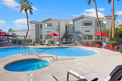 A sunny resort-style pool area here at Pala Mesa featuring a sparkling swimming pool with an attached spa, expansive concrete deck, and comfortable lounge seating beneath vibrant red umbrellas. Tall palm trees and manicured landscaping surround the courtyard, creating a relaxing outdoor retreat perfect for soaking up the Arizona sunshine.