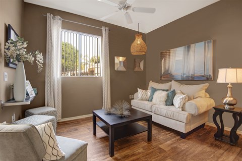 A cozy and stylish living room here at Pala Mesa featuring warm wood-style flooring, rich accent walls, and a plush cream sofa accented with textured throw pillows. A sleek dark coffee table anchors the space beneath a woven pendant light, while large windows with vertical blinds invite natural light and create a comfortable, inviting atmosphere.