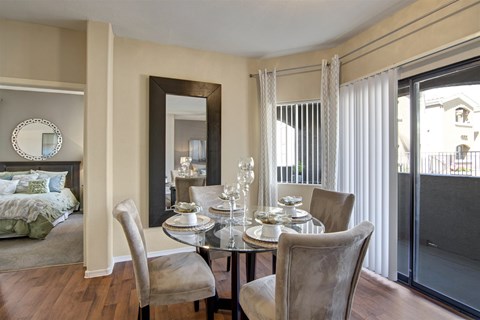 A bright dining area here at Pala Mesa featuring a round glass-top table with four upholstered chairs set on wood-style flooring. Large sliding glass doors with vertical blinds bring in natural light and lead to a private patio, while a full-length mirror enhances the open feel and reflects the adjacent bedroom entry.