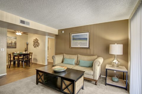 A warm living room here at Paradise Falls featuring a plush beige sofa with teal accent pillows, a dark wood coffee table, and soft carpeting, opening toward a dining area with wood-style flooring, coordinated décor, and overhead lighting, while neutral walls, framed artwork, and a table lamp create a comfortable and inviting atmosphere.