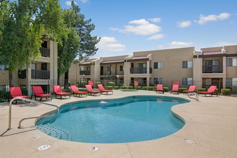 A resort-style swimming pool here at Paradise Falls featuring a curved pool with clear blue water, surrounding concrete deck, and a row of red lounge chairs along a black metal fence, with mature trees and tan apartment buildings in the background creating a relaxing outdoor setting under open skies.
